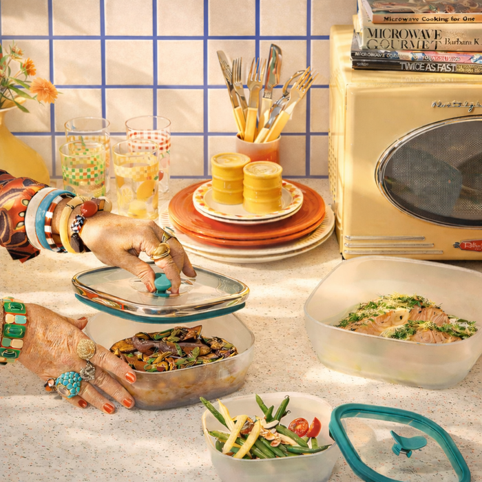 A person with colorful jewelry opens a container from the 4-Piece Glass Square Dish Set on a kitchen counter, with plates, glasses, utensils, an old microwave, and cookbooks visible in the background.