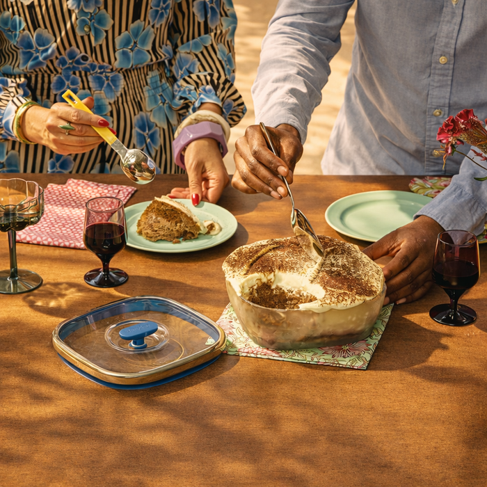 Two people serve tiramisu from a 10-Cup Glass Square Shallow Dish onto plates at an outdoor table, surrounded by wine glasses, colorful napkins, and a flower vase. The scene feels warm and inviting.