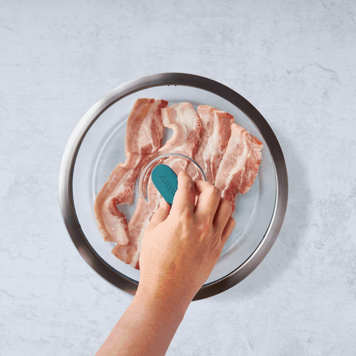 A hand presses a 12-Cup Glass Round Deep Dish over slices of raw bacon arranged in a circle on a round glass plate, set on a light gray surface—an efficient meal-prepping step perfect for feeding a crowd or preparing big batch soups.