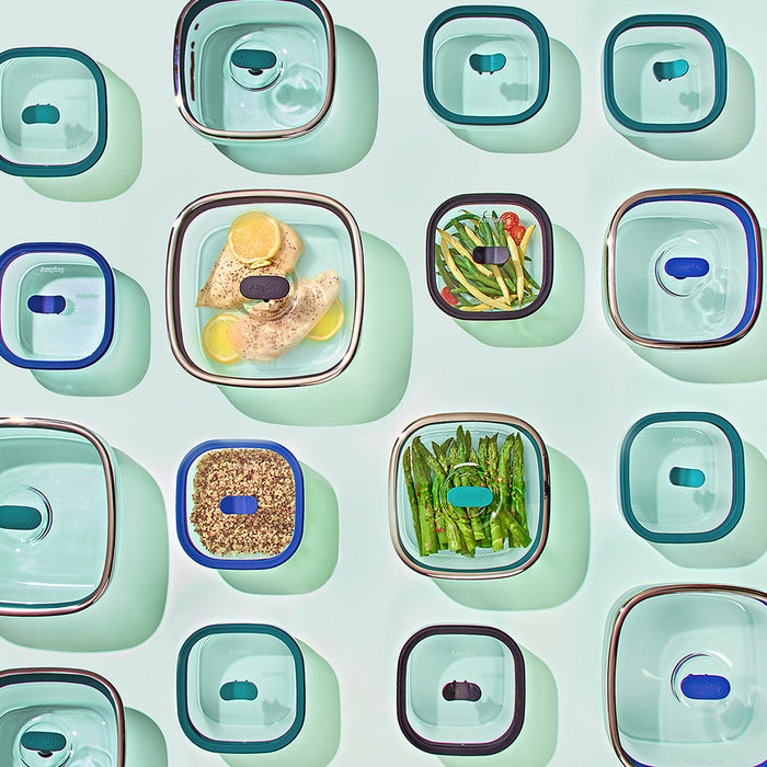 Four food storage containers from the 8-Piece Glass Square Dish Set, filled with chicken with lemon, vegetables, and grains, are arranged alongside empty dishes and lids in a neat grid on a pale green surface. The frosted glass of the cookware lends an elegant touch to this colorful and organized display.