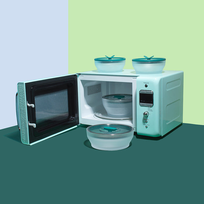 An open mint green microwave reveals a covered container from the 8-Piece Glass Round Dish Small Spaces Set inside, while three additional matching containers from the set—two stacked on top and one placed in front—are displayed against an eye-catching two-tone green and blue backdrop.