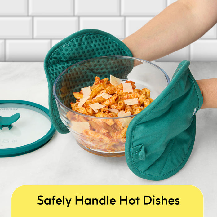 A person uses green oven mitts to hold a glass bowl of pasta with sauce and cheese, featuring the Anyday Accessory Bundle—perfect for microwave cooking. A matching green lid from the handy storage accessories rests nearby on the white countertop. Text below reads, “Safely Handle Hot Dishes.” White tiled wall in background.