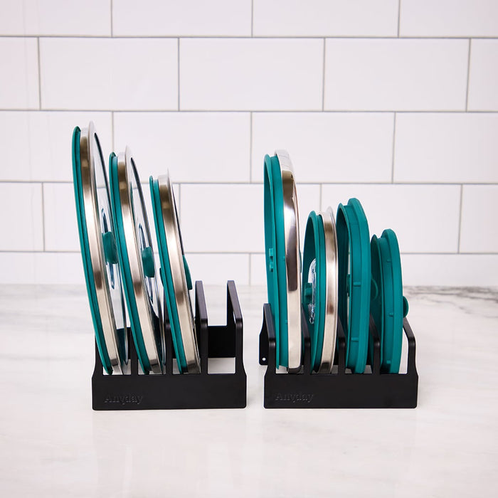 Two Lid Organizer racks in black hold several teal and silver pot lids upright on a white marble countertop, set against a white tiled wall.