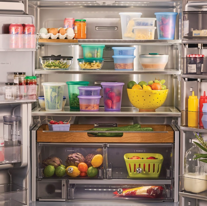 Open refrigerator filled with various foods, including eggs, canned drinks, fresh fruits, vegetables in drawers, sauces, and produce neatly stored in reusable Anydeli Mixed Set platinum silicone containers (6-pack), with a bottle of milk on the door shelf. Items are beautifully organized and colorful for efficient food storage.