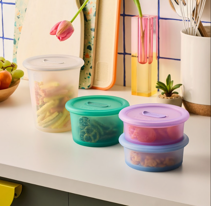 Six vibrant Anydeli Mixed Set reusable plastic food containers with lids, filled with fruits and vegetables, are arranged on a white kitchen counter. Cutting boards, a pink tulip in a vase, and kitchen utensils can be seen in the background—ideal for keeping your food organized all week long.