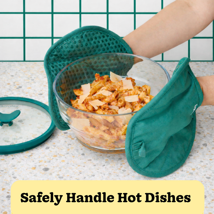 Wearing Anyhand Mitts in teal, a person securely holds a glass bowl of hot pasta. The bowl rests on heat-resistant trivets atop a speckled countertop, with a glass lid set nearby. Text below reads: Safely Handle Hot Dishes.