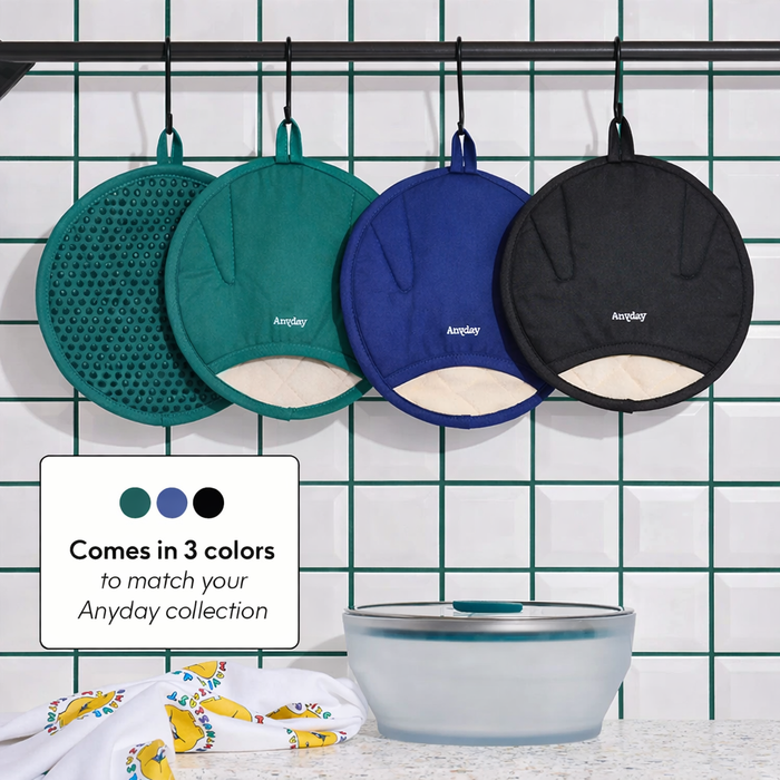 Four round, heat-resistant Anyhand Mitts in green, blue, and black hang on hooks against a tiled wall; text reads: Comes in 3 colors to match your Anyday collection. Designed to fit any hand comfortably. A clear glass bowl and a colorful towel are below.