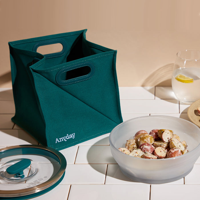 A teal fabric tote labeled Anywhere Bag sits next to a glass dish of seasoned potatoes—part of the versatile Anyday dishes collection—on a white tiled surface, with a lid nearby and a glass of water with lemon in the background, highlighting the convenience of this microwave cookware and portable food container for everyday meals.