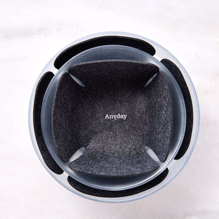 Top-down view of the Dish Protector, a round container with four rectangular side openings and a dark, felt-like liner labeled Anyday inside, designed for scratch-free storage. The container is placed on a light, textured surface.