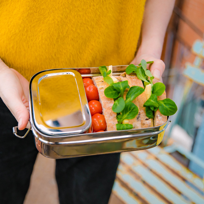Stainless steel lunch box with snack pod dividing main compartment of the lunch box.showing a sandwich and tomatoes