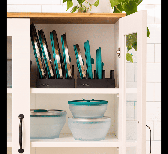 A kitchen cabinet with glass doors displays a Lid Organizer neatly keeping lids upright on the top shelf, while three frosted mixing bowls with teal lids are arranged on the lower shelf. A green plant hangs above.