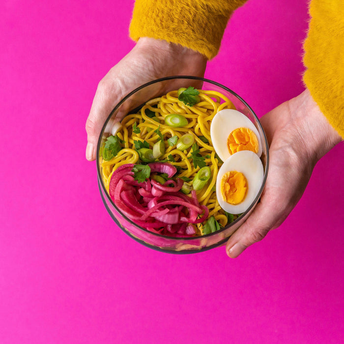 elephant box glass lunch bowl on pink background with a meal of noodles and egg.