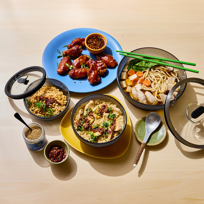 Four dishes on a table—noodles with chicken and vegetables, herbed rice, glazed chicken wings on a blue plate, and fried rice topped with sauce—each elegantly presented in dishes from the Onyx Collection. Also featured are utensils, sauces, green chopsticks, and a selection of microwave meals.
