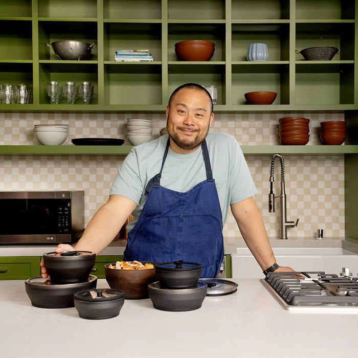 Wearing a blue apron and standing in a modern kitchen with green shelves, a person smiles as they prepare food, surrounded by dishes from the Onyx Collection—demonstrating that microwave meals can be served with style.