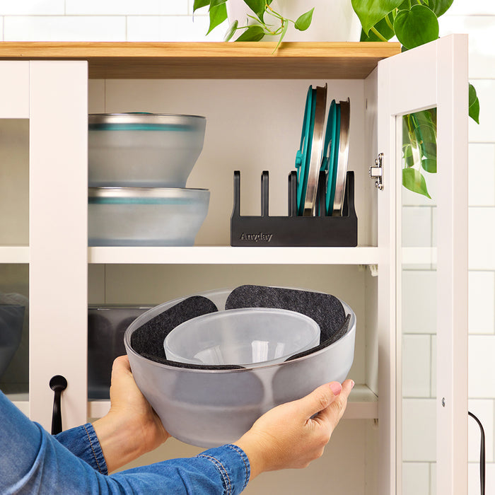 A person stacks ceramic bowls with felt protectors in a kitchen cabinet, showcasing thoughtful cabinet organization using the Storage Bundle. Cookware lids stand vertically in a black organizer on the shelf above, next to more bowls and Anyday Dishes accessories. Green plant leaves hang above the cabinet.