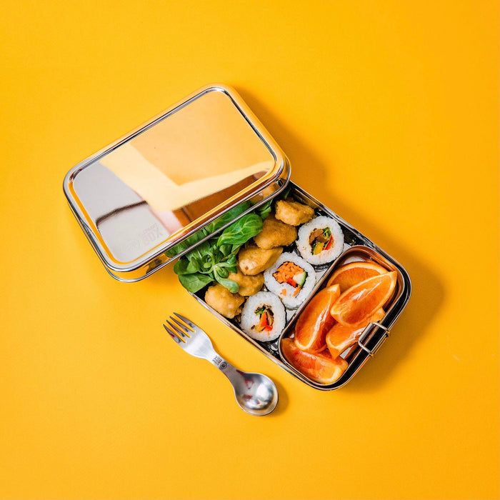 a rectangular stainless steel lunchbox on a bright yellow background, the main compartment of the lunchbox contains sushi and the removable snack pod contains pieces of fruit. A stainless steel spork is laid to one side.